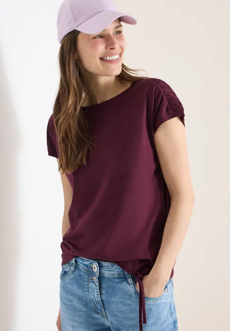Frau trägt lila T-Shirt mit Knoten am Saum und Jeans, vor hellem Hintergrund.